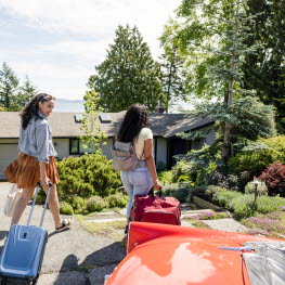Två unga kvinnor med resväskor går mot ett hus omringat av träd och med utsikt över vatten.