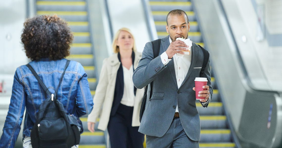 Man in suit eating a sandwich and holding a take away cup while walking from escalators, with other people around him.