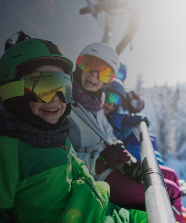 Familj i skidutrustning sitter i en sittlift och ler mot kameran.