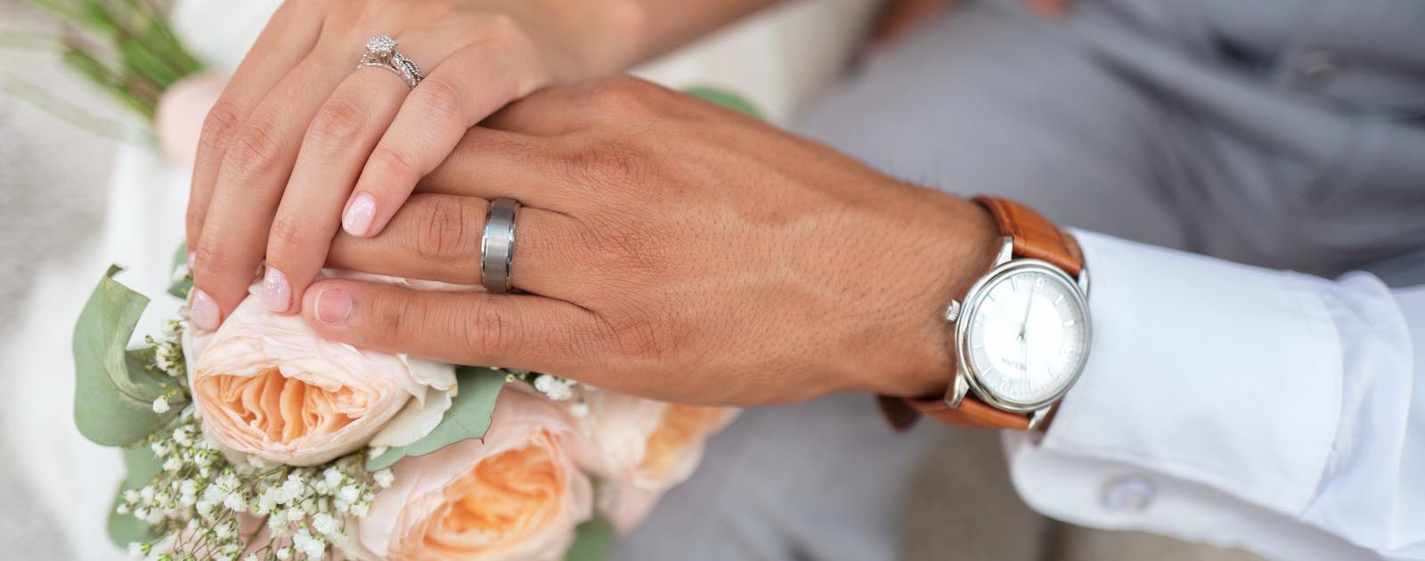 Female hand with wedding band over male hand with wedding band, both on top of a bouquet.