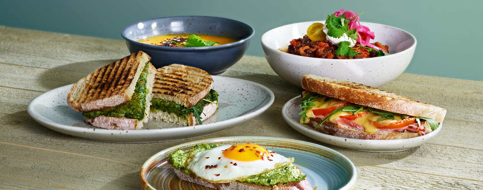 Three plates, each with a sandwich on it and two bowls in the back, one containing soup and one containing grilled vegatables.