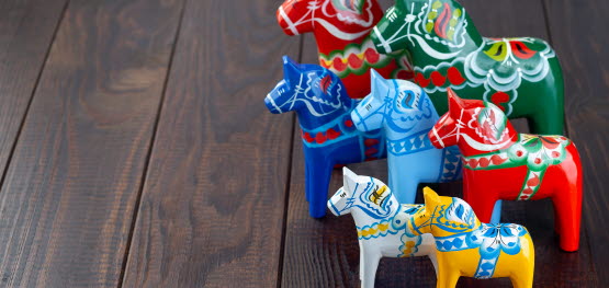7 dala horses in different sizes and colours on a wooden floor.