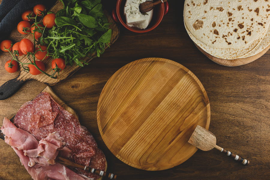 Tortilla, salami, skinka, tomater, ruccola, färskors och en träbricka på ett bord.