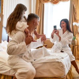 Family clad in robes in hotel room, mom taking picture of dad and daughter who is sitting on his shoulders.