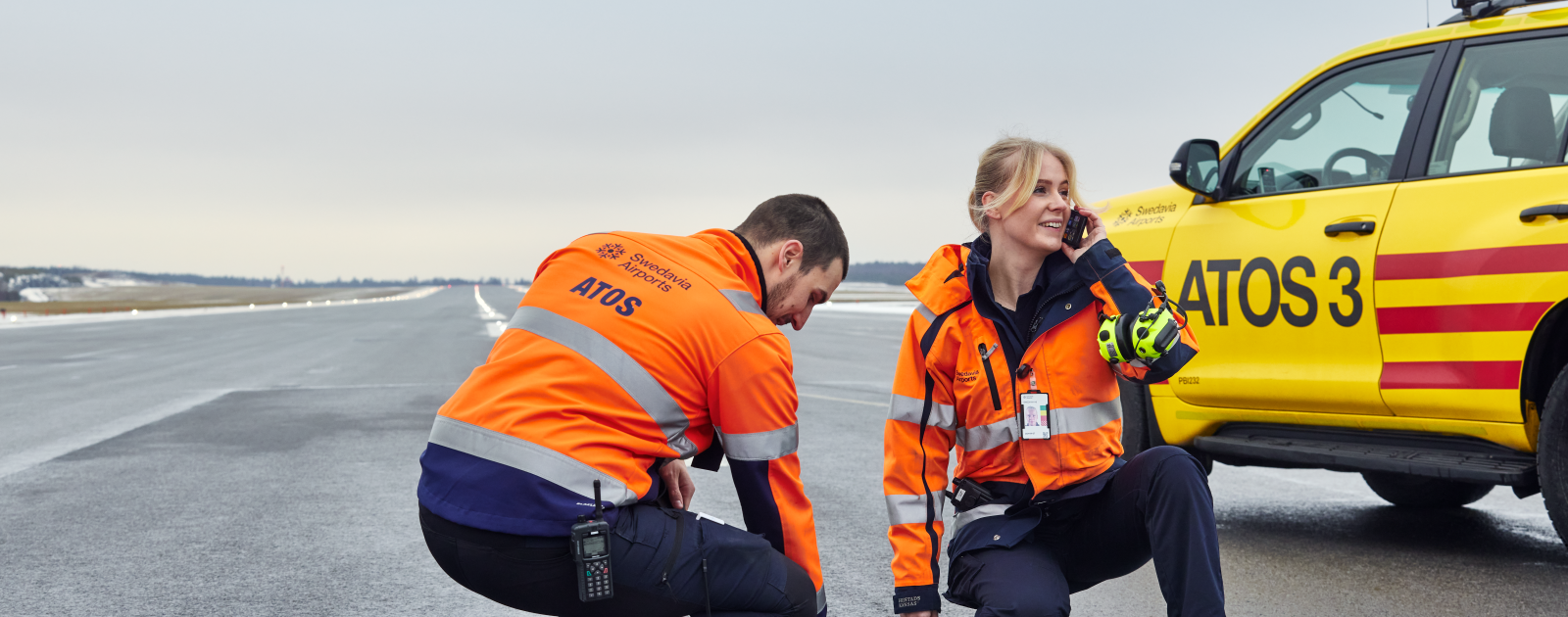 Två ATOS-medarbetare på landningsbanan, en petar på ett banljus medan kvinnan pratar i telefon.