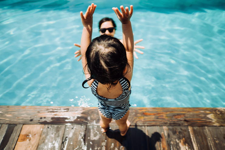 Little girl in swimsuit standing with arms up by the poolside, with mom in water ready to catch her.