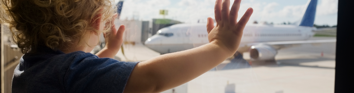 Toddler standing at a window looking at an airplane.