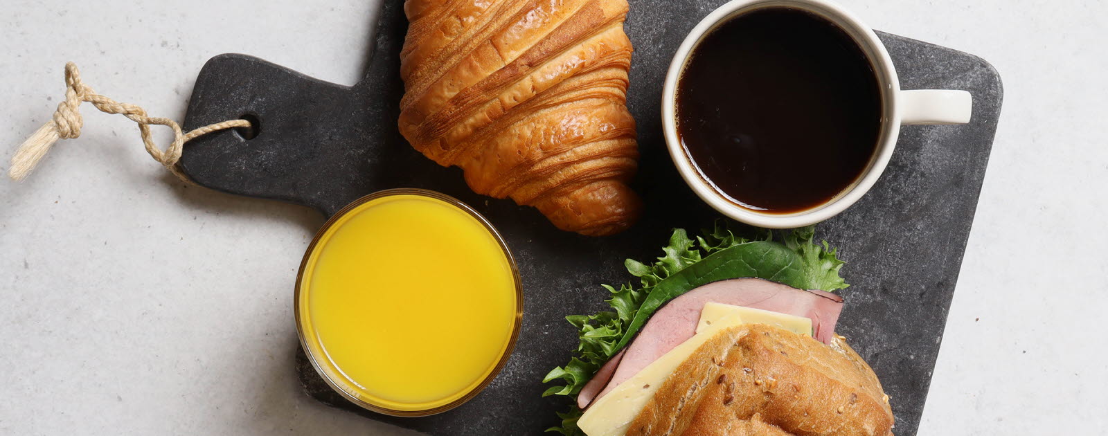 Grå marmorbricka med en ost- och skinkfralla, en croissant, en apelsinjuice och en svart kaffe.