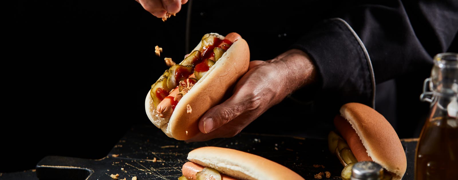 Man's hands preparing hotdogs with pickles, ketchup and roasted onion.