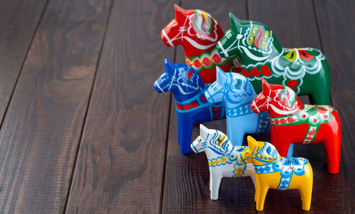 7 dala horses in different sizes and colours on a wooden floor.