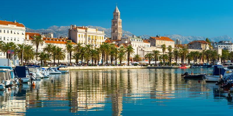 Harbour with motorboats, a boardwalk lined with palms, Mediterranean style buildings and a church tower, all reflected in the water.