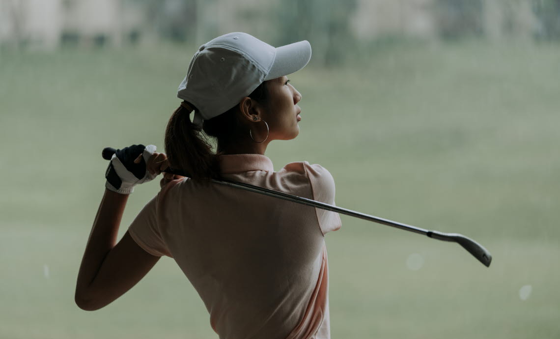 Young woman in white hat taking a golf swing.