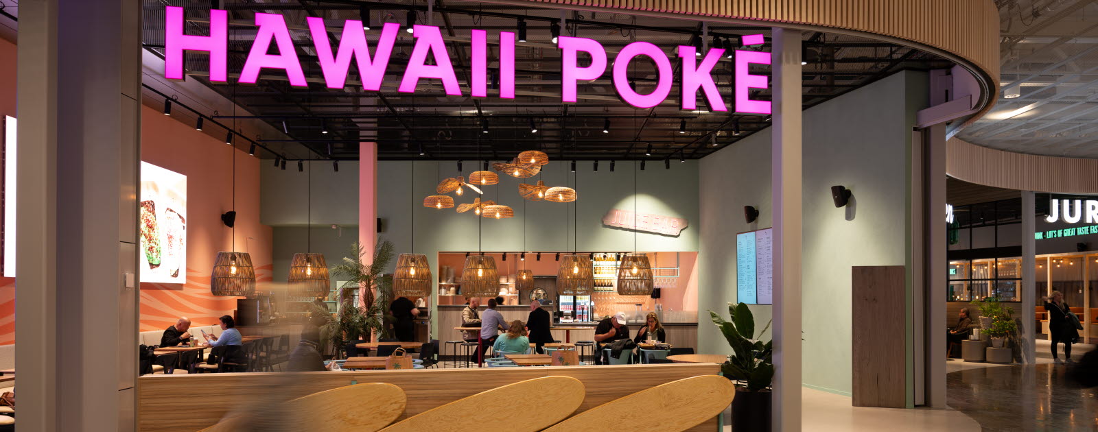 Restaurang med turkos- och korallfärgad vägg, surfinspirerad inredning och en stor rosa neonskylt med texten "Hawaii Poké".