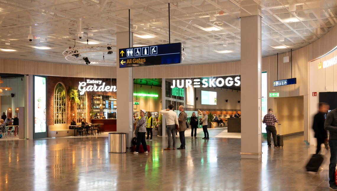 People with suitcases standing and walking in an open area in front of restaurants and shops.