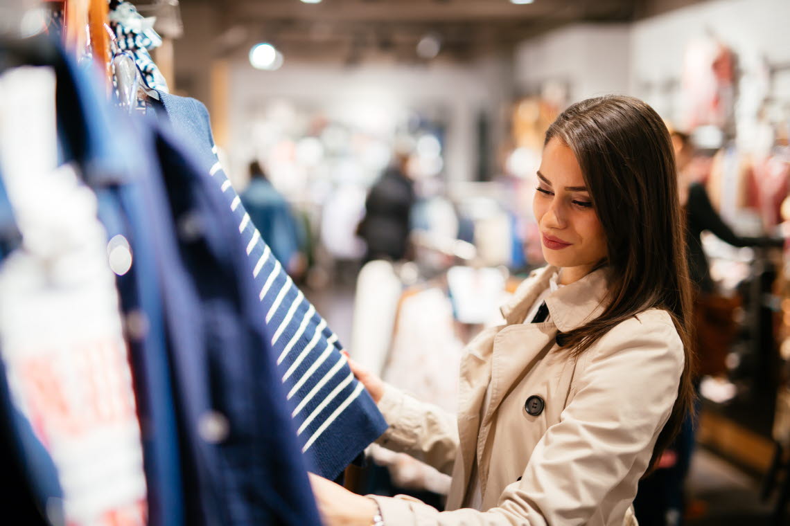 Woman with long brown hair dressed in a trenchcoat looking at a striped sweater.