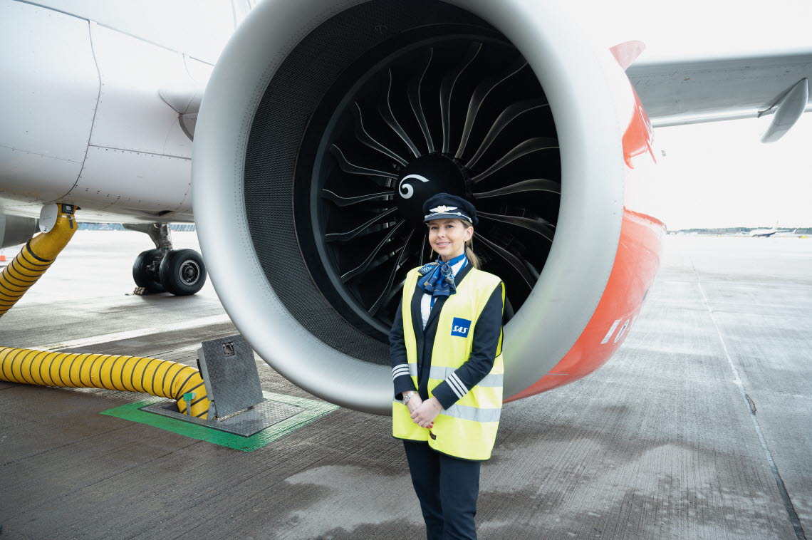 Female pilot in high visibility vest standing in front of jet engine.