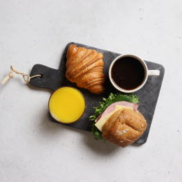 Grå marmorbricka med en ost- och skinkfralla, en croissant, en apelsinjuice och en svart kaffe.