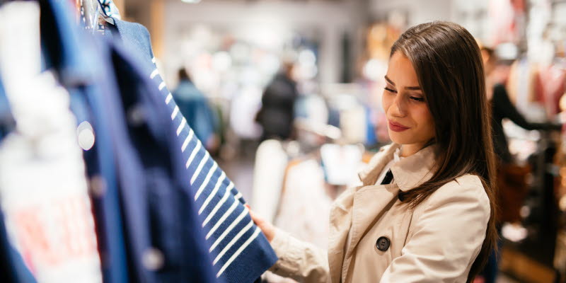 Woman with long brown hair dressed in a trenchcoat looking at a striped sweater.