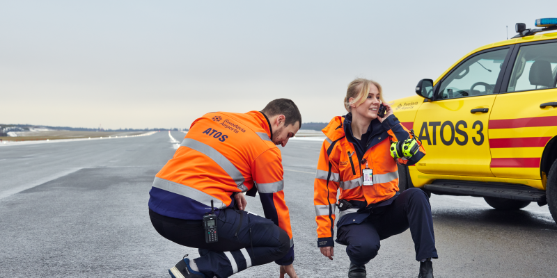 Two ATOS staff on the runway, one poking at a runway light while the female staff is talking on the phone.