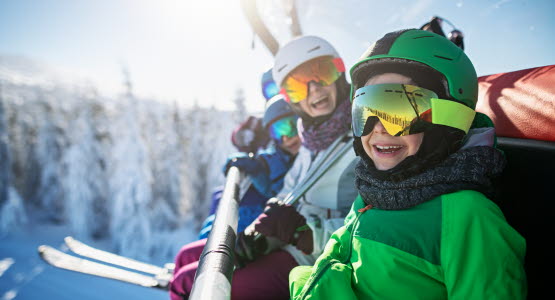 Family in ski equipment sitting in chair lift smiling at the camera.