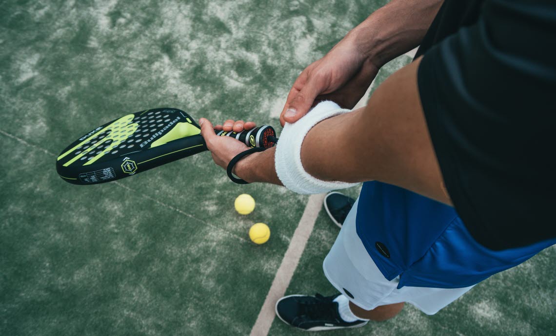 Person dressed in workout gear, putting a sweat band on one arm while holding padel racket