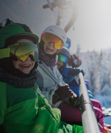 Familj i skidutrustning sitter i en sittlift och ler mot kameran.