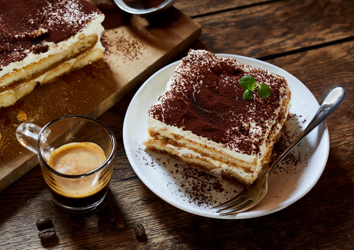 A piece of tiramisiu and a fork on a plate next to an espresso.