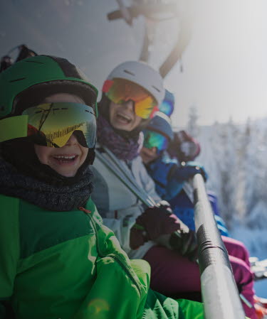 Familj i skidutrustning sitter i en sittlift och ler mot kameran.