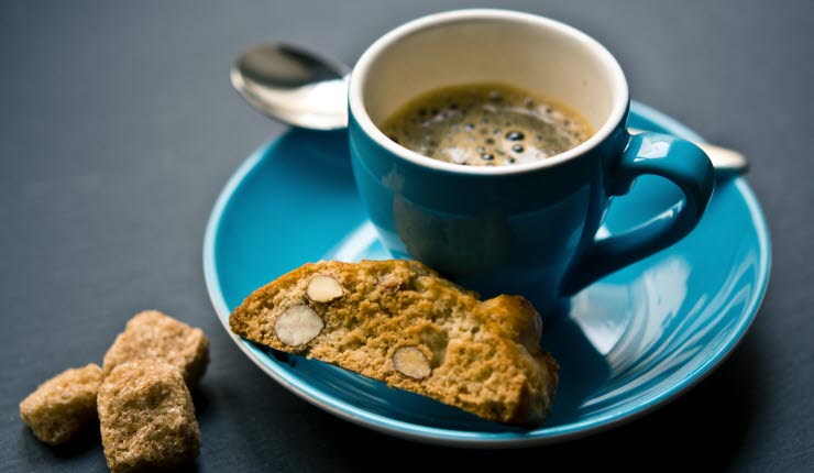 En espresso i blå kopp med biscotti och en sked på kaffefatet.