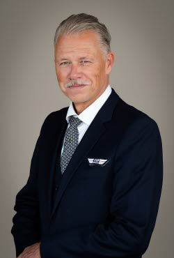 Man with slicked back hair and moustache dressed in a suit and tie smiling at the camera.