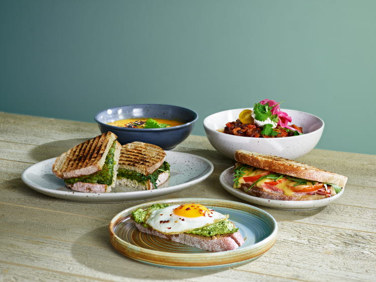 Three plates, each with a sandwich on it and two bowls in the back, one containing soup and one containing grilled vegatables.