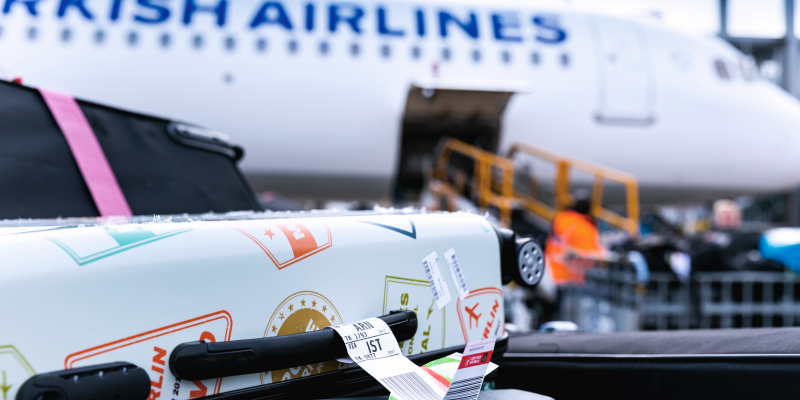 Hörn av två resväskor med bagagetaggar framför ett flygplan som lastas.