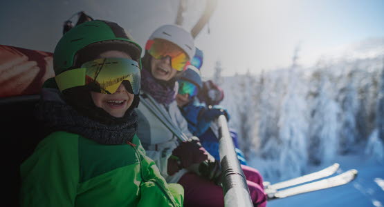 Familj i skidutrustning sitter i en sittlift och ler mot kameran.